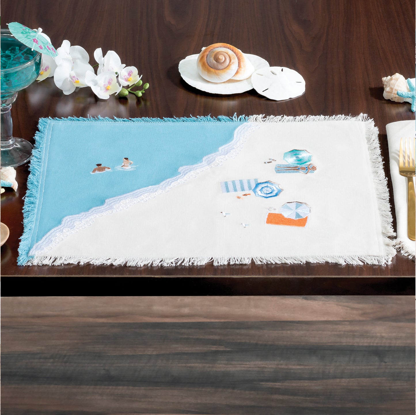 Colorful beach umbrellas, towels, and sun bathers embroidered on a sandy beach scene of natural and blue cotton on a fringed placemat.