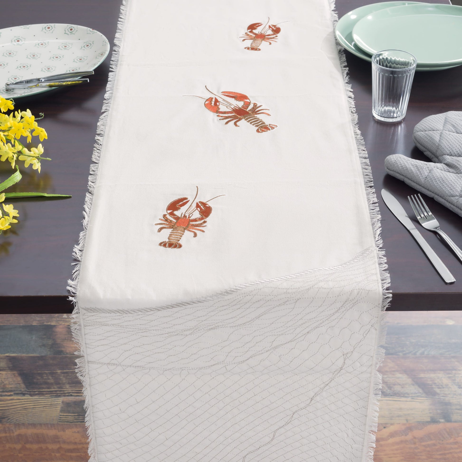 Natural fringed cotton table runner featuring embroidered red lobsters set on table with dishes.