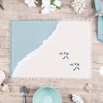 Two sandpipers embroidered on a cotton fringed placemat featuring blue waves on sand. Placemat is sitting on a wooden table.