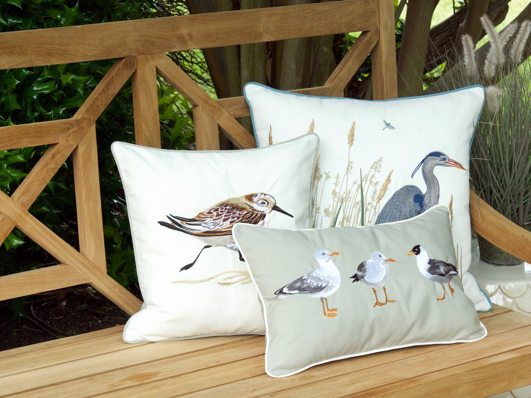Three pillows on an outdoor bench each embroidered with sea birds. Pillow one has 3 sea gulls, pillow 2 has an avocet, and pillow 3 has a heron.