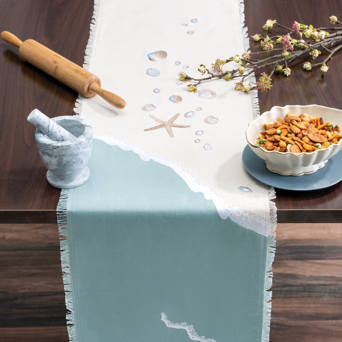 Sea stars and sea shells embroidered on a cotton runner featuring a wave washing onto the beach, finished with fringe edges on a table with snacks and cooking tools.