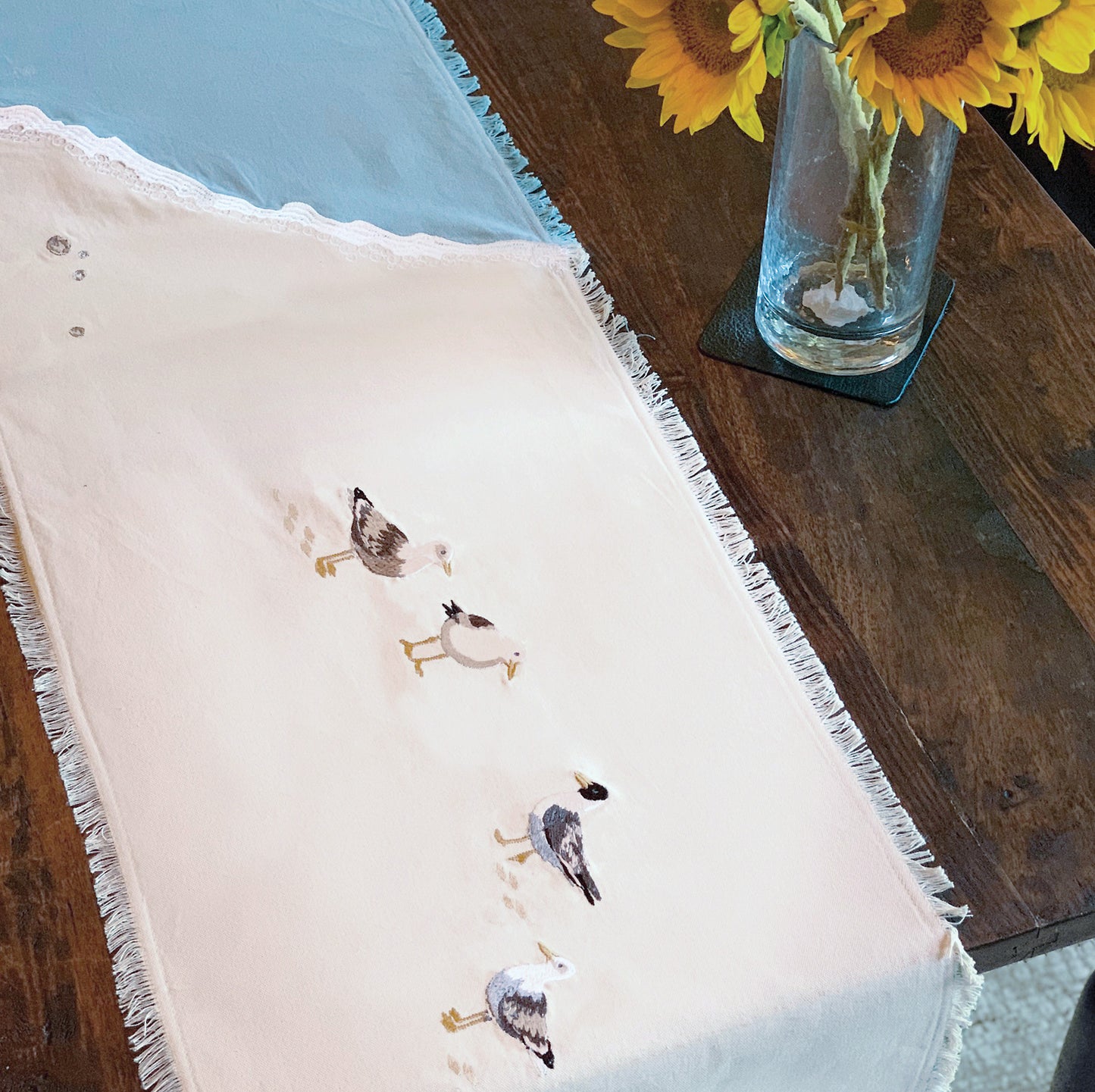 Sea gulls embroidered on a cotton fringed table runner featuring blue waves on sand. Linens are sitting on a wooden table.