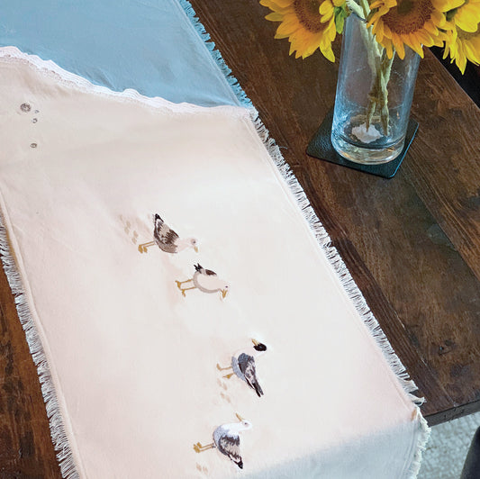Sea gulls embroidered on a cotton fringed table runner featuring blue waves on sand. Linens are sitting on a wooden table.