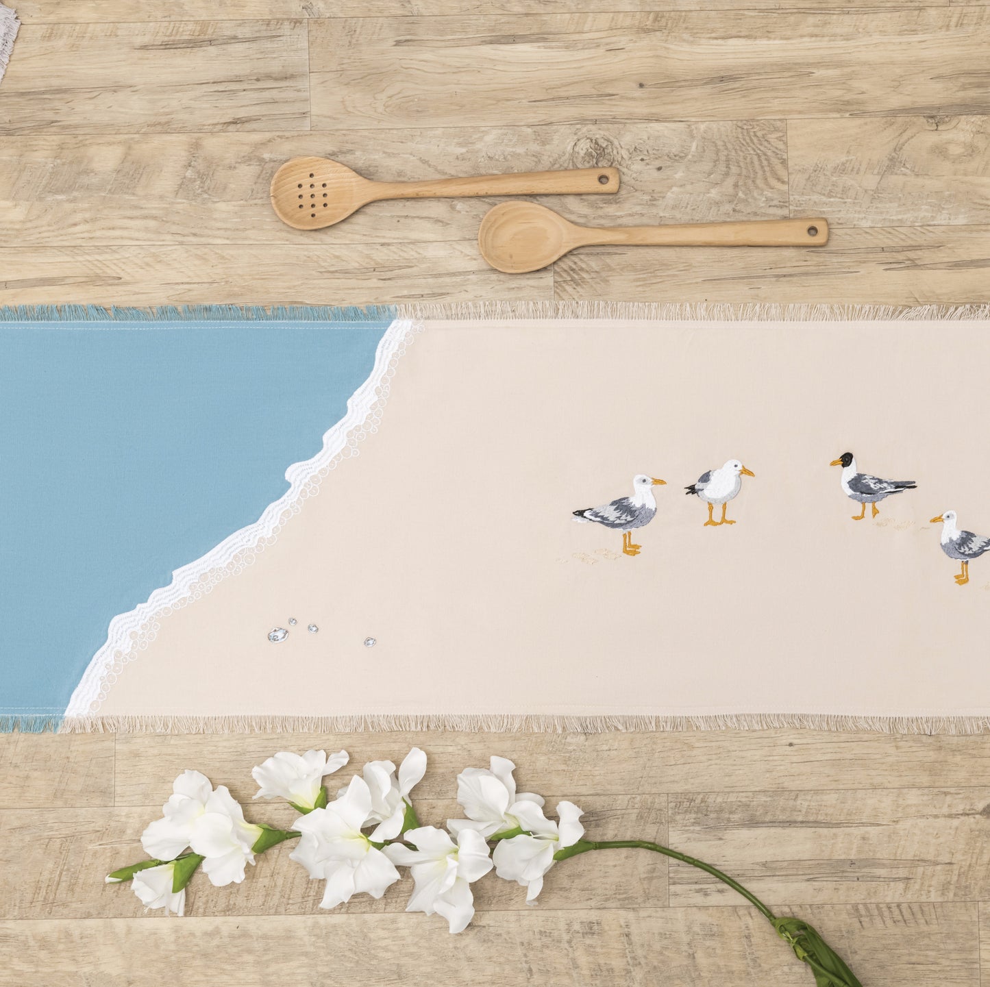 Sea gulls embroidered on a cotton fringed table runner featuring blue waves on sand. Linens are sitting on a wooden table.