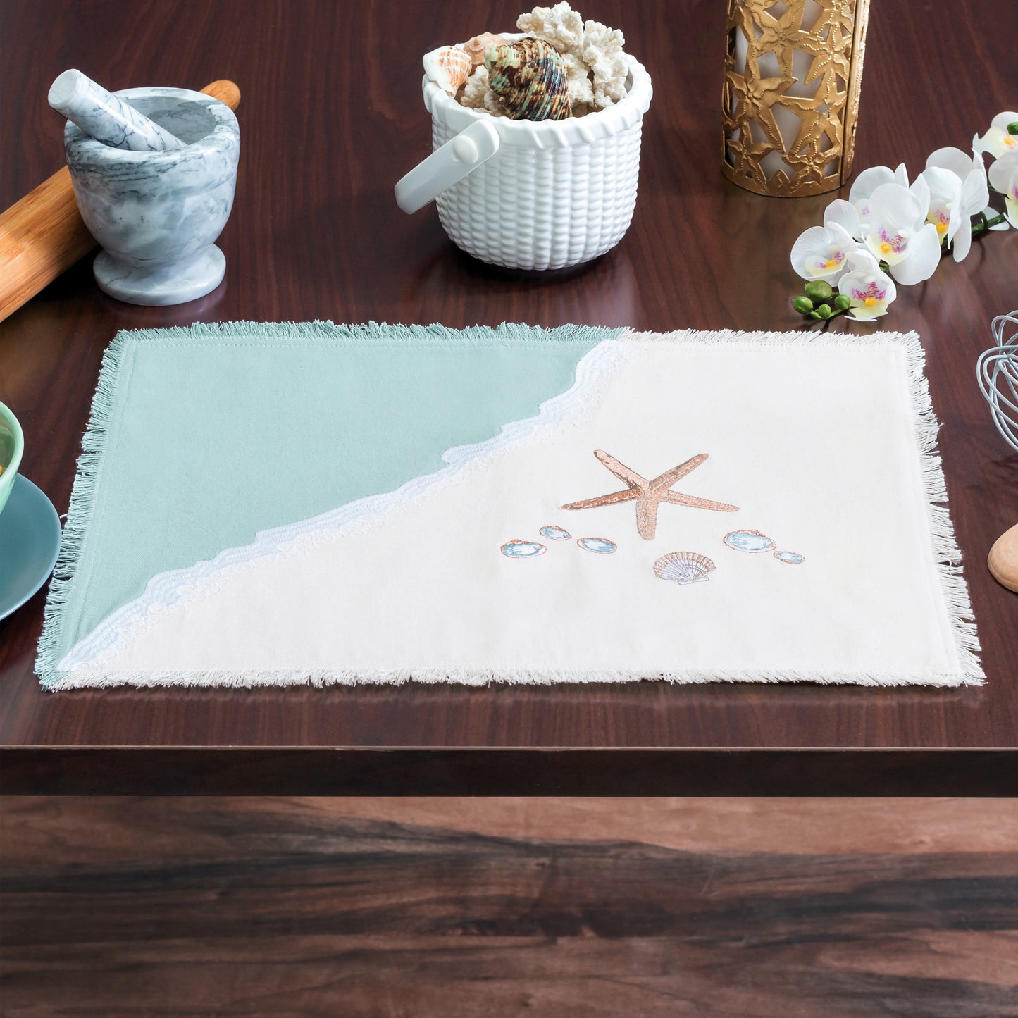 Sea stars and sea shells embroidered on a cotton and placemat featuring a wave washing onto the beach, finished with fringe edges SET ONA WOODEN TALBE WITH ACCESSORIES.