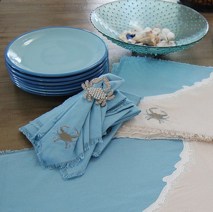 Baby blue crabs embroidered on sand colored ground and blue wave with embroidered sea foam placemat and runner. Blue crab embroidered on a coastal blue cotton napkin with fringe. On a table with dishes.