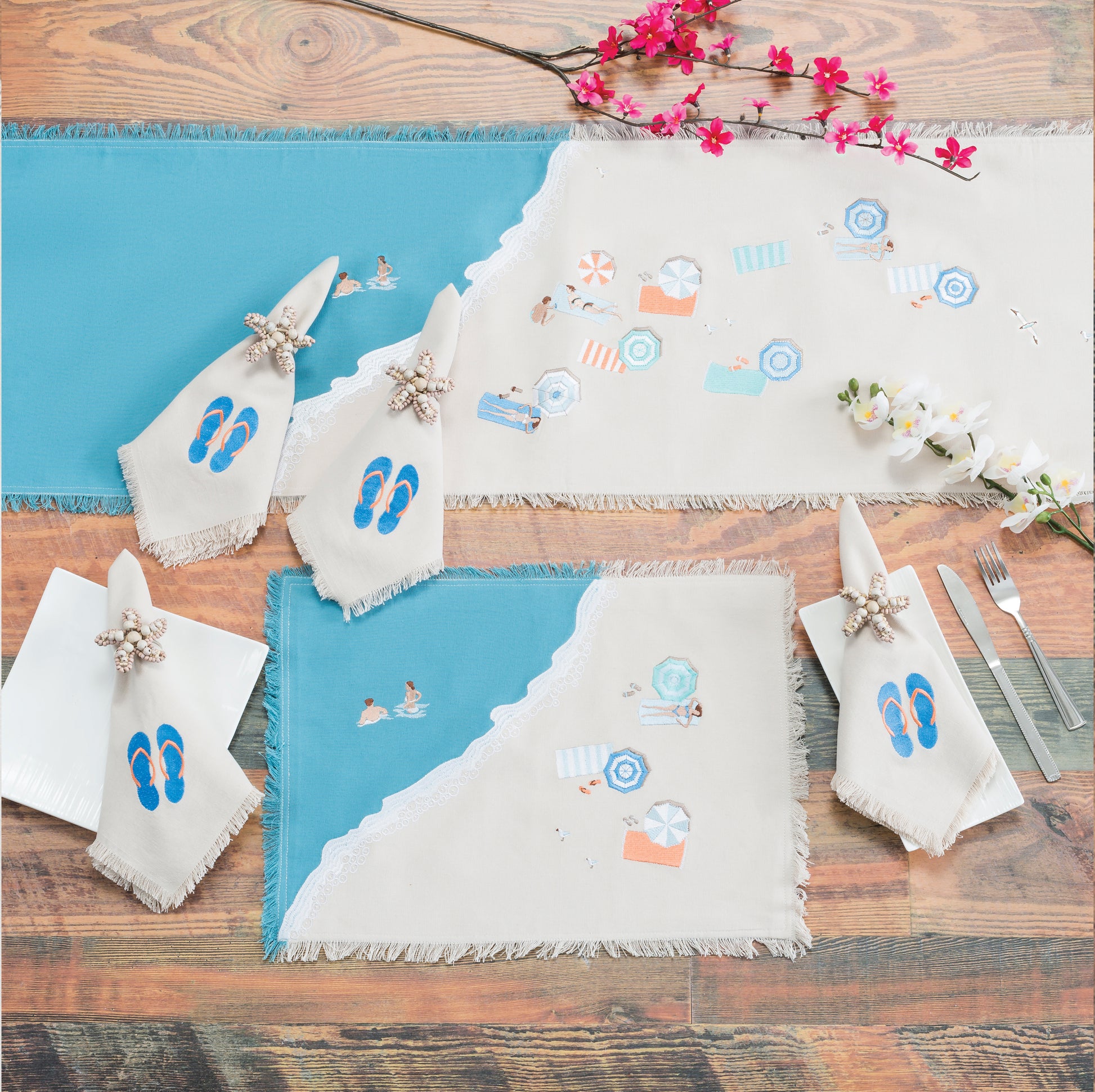 Colorful beach umbrellas, towels, and sun bathers embroidered on a sandy beach scene of natural and blue cotton on a fringed table runner and placemat. Napkins featuring embroidered flip flops not available. 