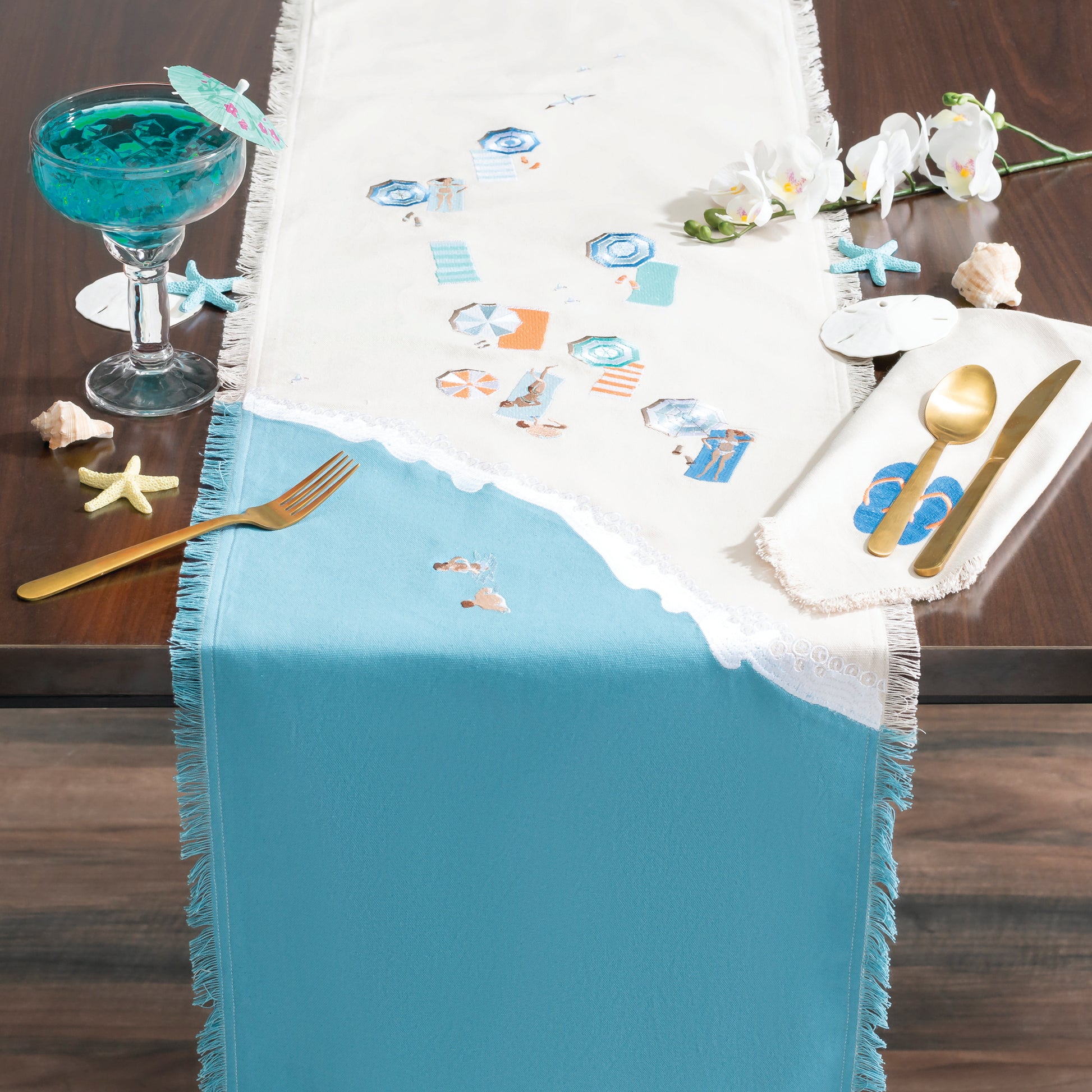 Colorful beach umbrellas, towels, and sun bathers embroidered on a sandy beach scene of natural and blue cotton on a fringed table runner.