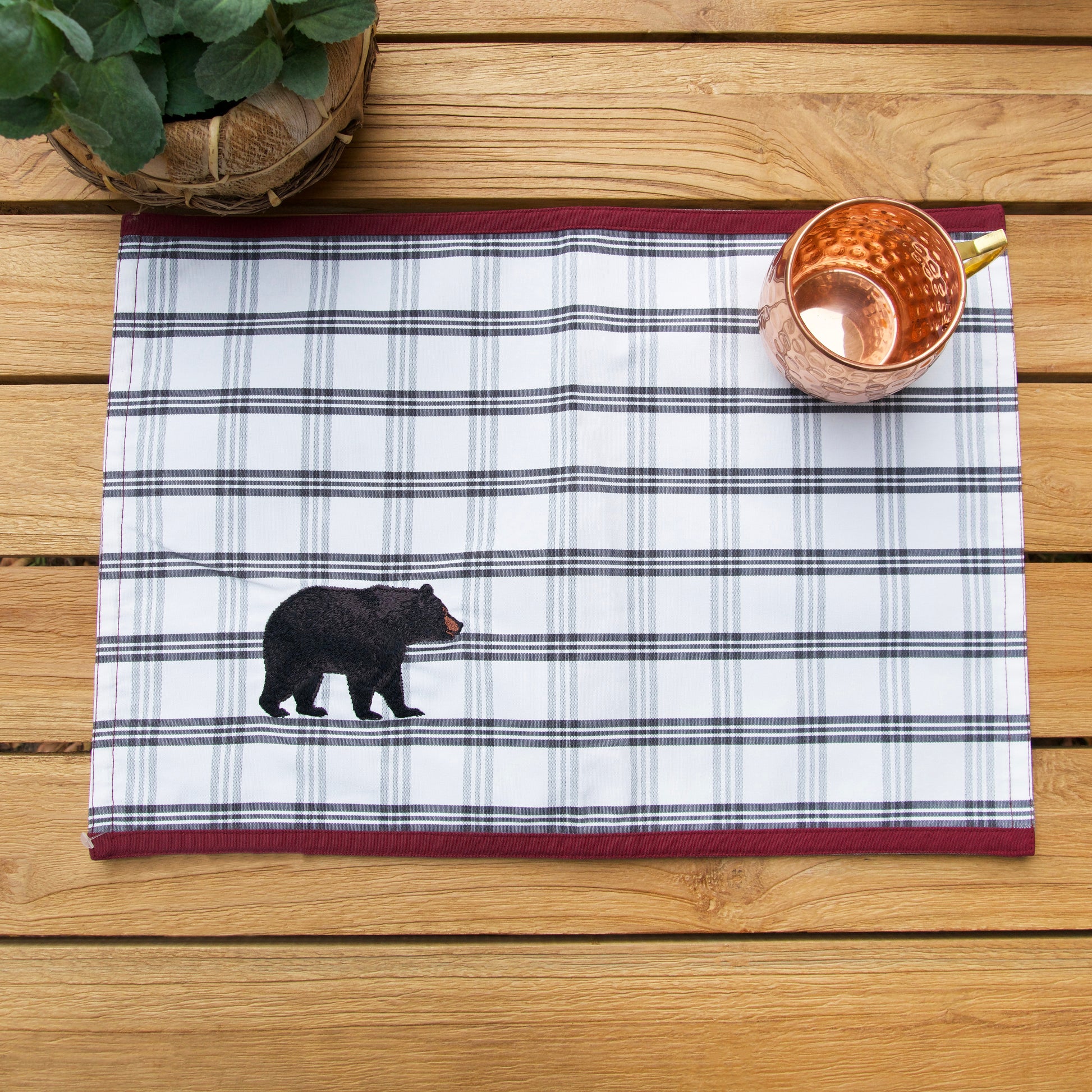 White placemat with grey plaid and a black bear in the right corner featuring a red border on the top and bottom on an outdoor table