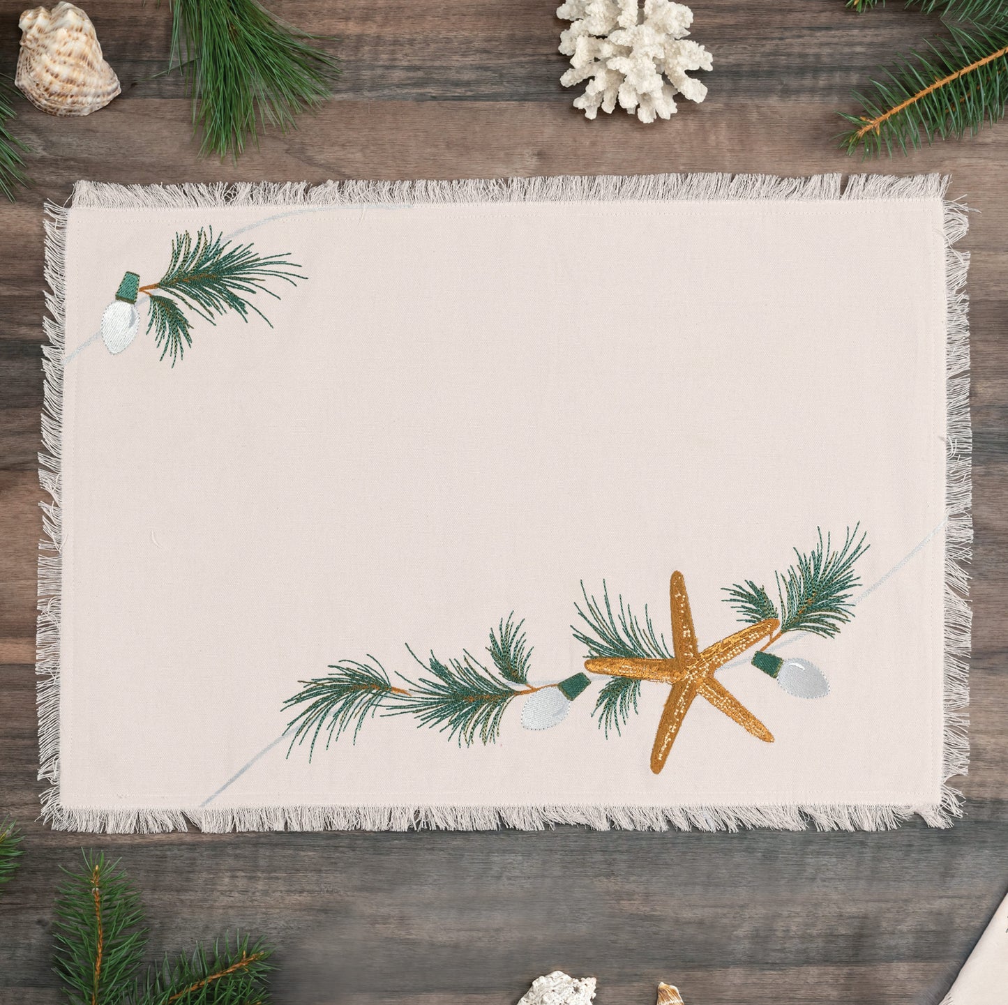 Natural cotton fringed placemat featuring an embroidered sea star, holiday lights, and evergreen needles laid on a wooden table.