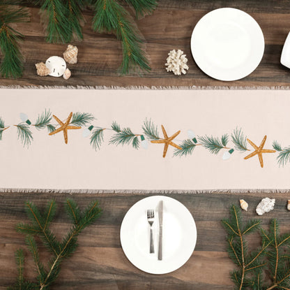 Natural cotton fringed runner featuring an embroidered sea stars, holiday lights, and evergreen needles laid on a wooden table with white dishes.