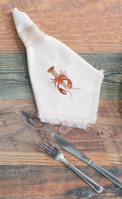 Natural fringed cotton napkin featuring embroidered red lobsters set on table with flatware.
