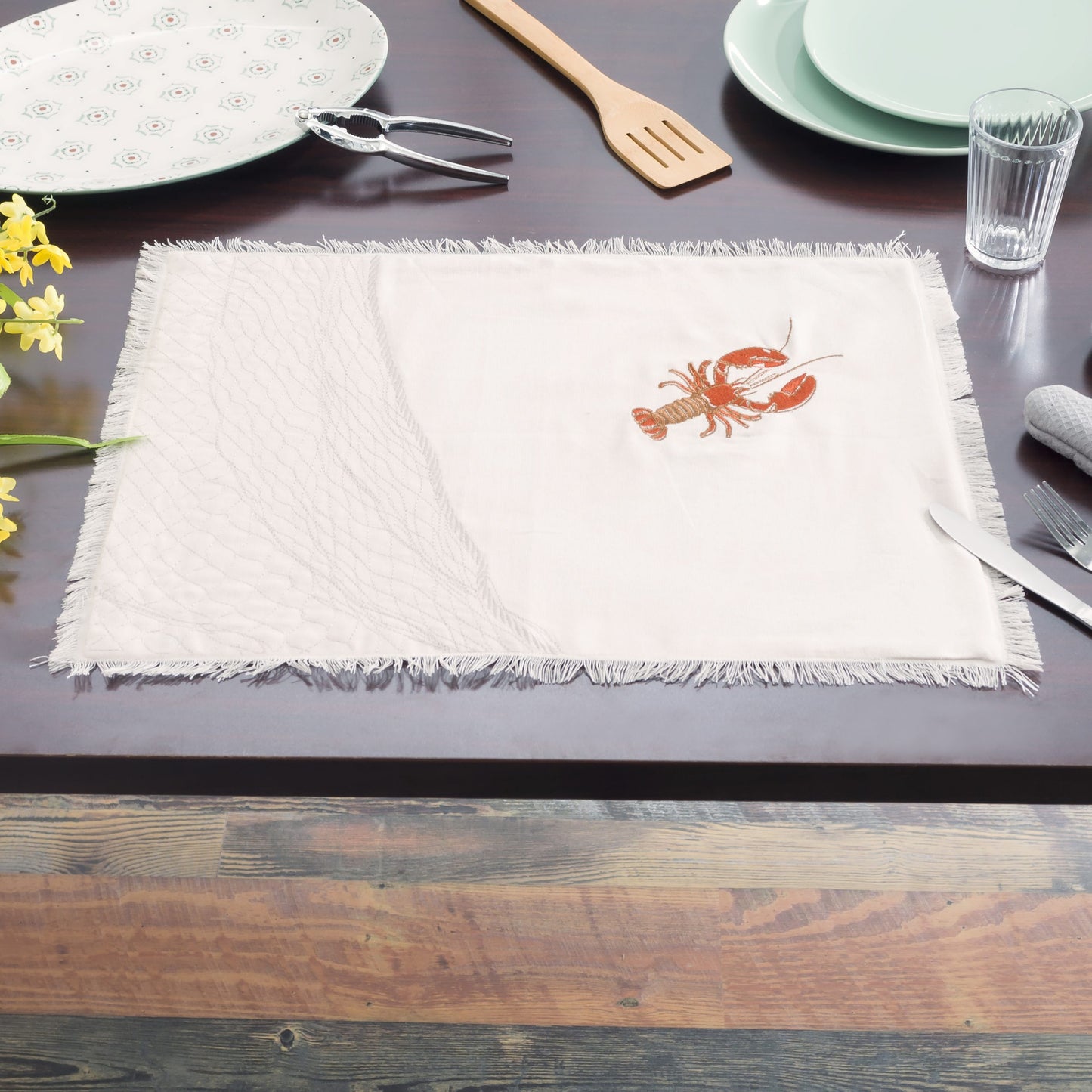 Natural fringed cotton placemat featuring embroidered red lobsters set on table with dishes.