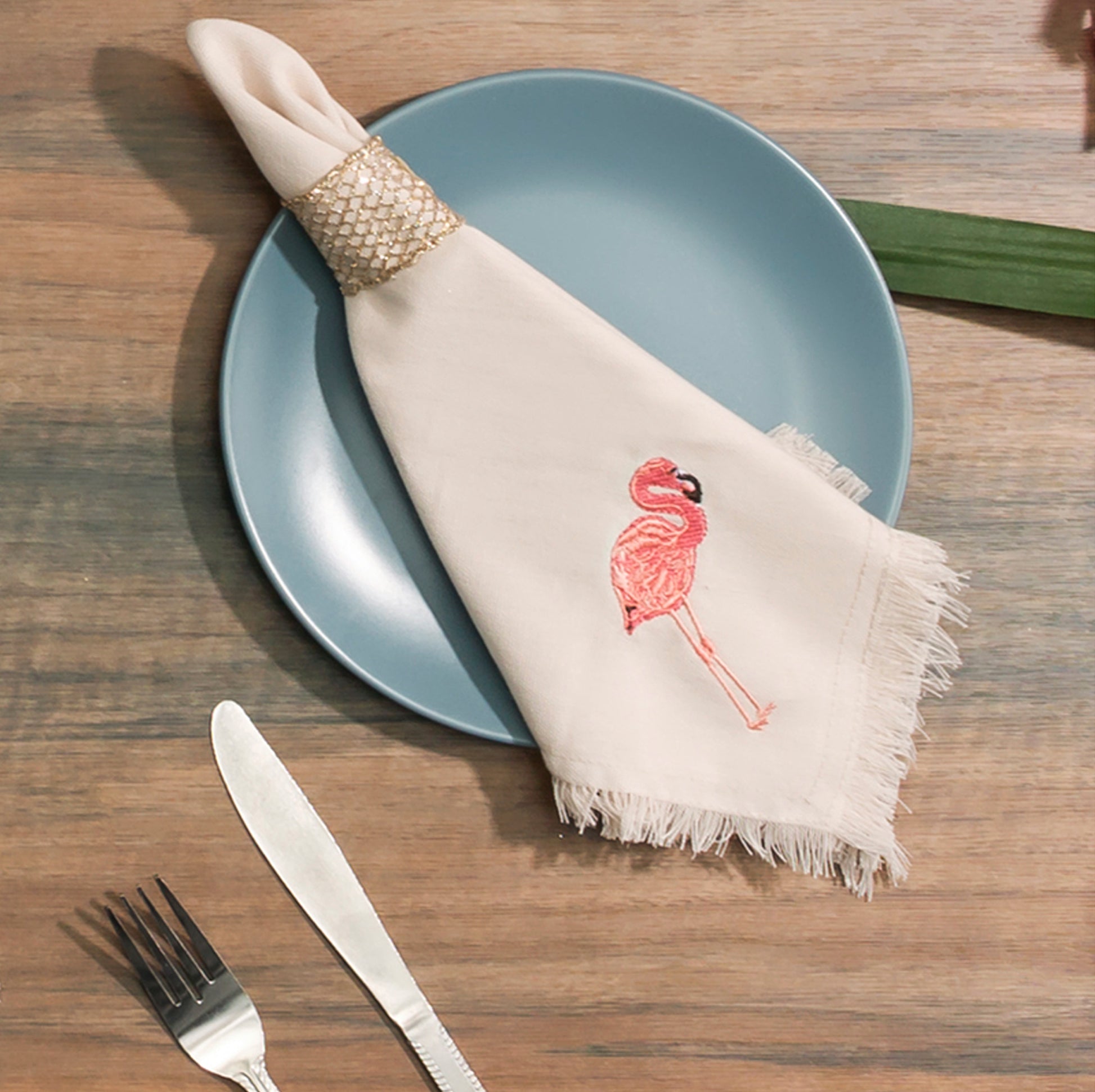 Napkin depicting embroidered Pink Flamingo on a tan cotton background with fringe edges. Napkin sitting on blue plate.
