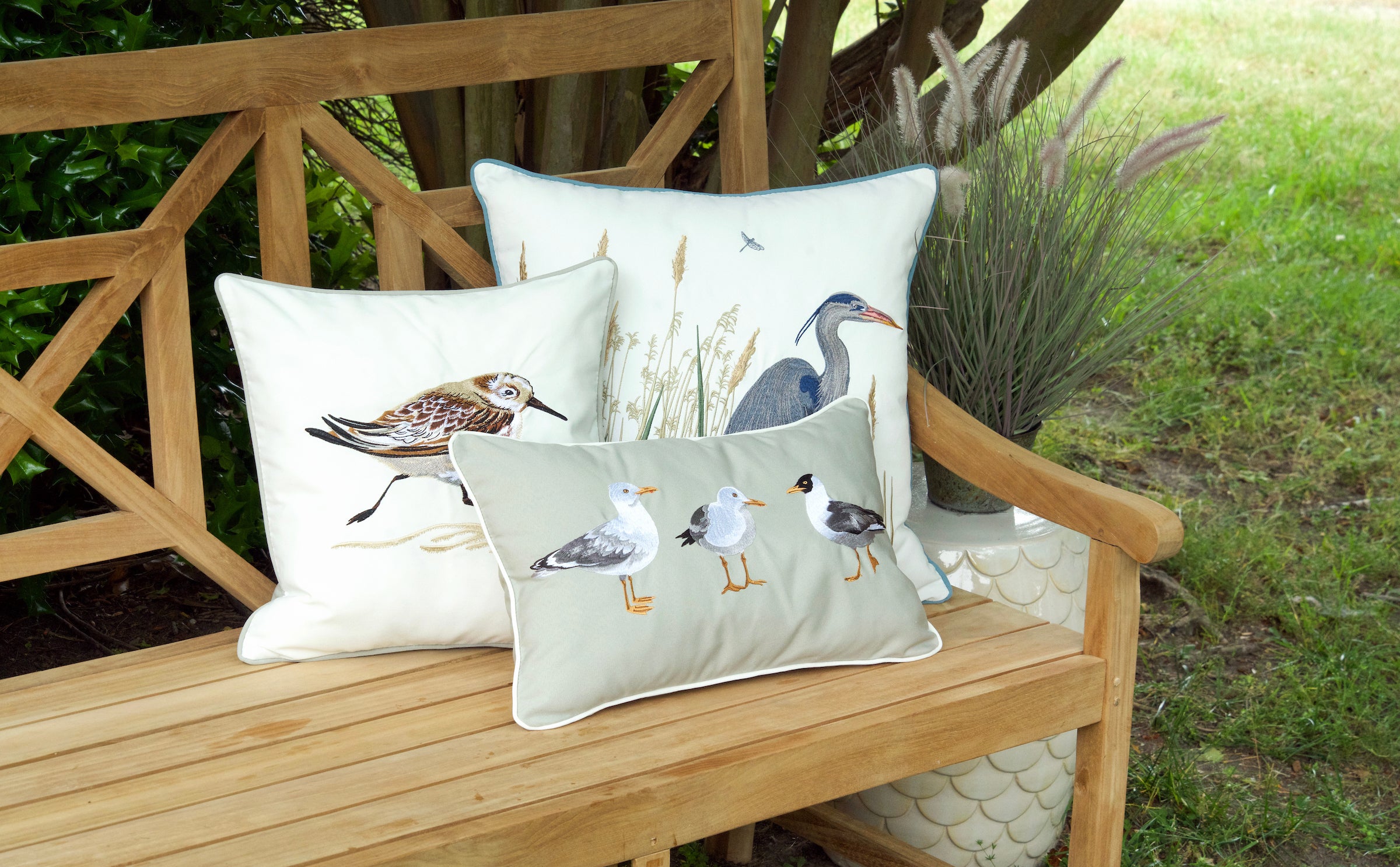 Three pillows on an outdoor bench each embroidered with sea birds. Pillow one has 3 sea gulls, pillow 2 has an avocet, and pillow 3 has a heron. 