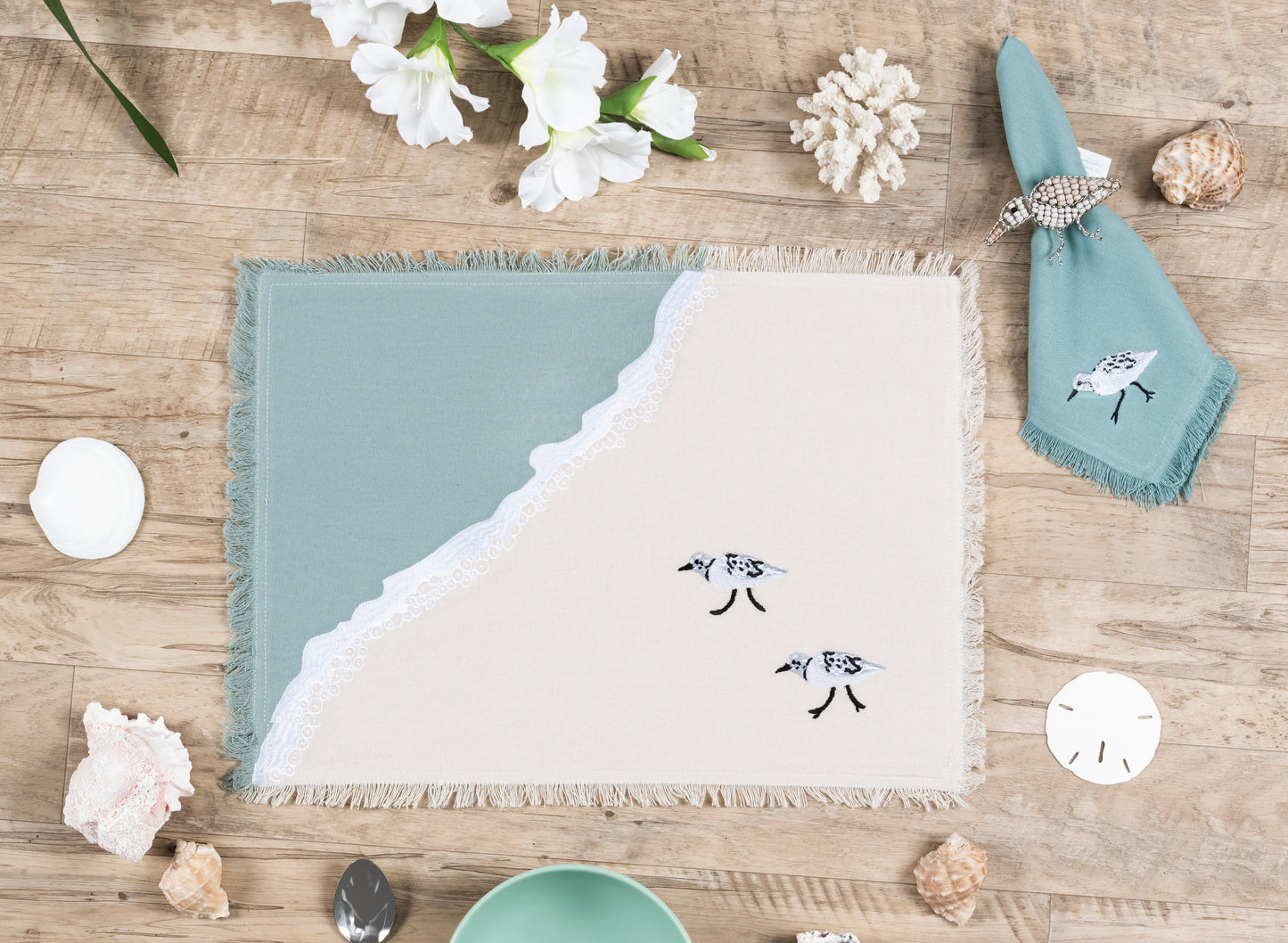 Sandpiper embroidered on coastal blue napkin next to two sandpipers embroidered on a cotton fringed placemat featuring blue waves on sand. Linens are sitting on a wooden table.