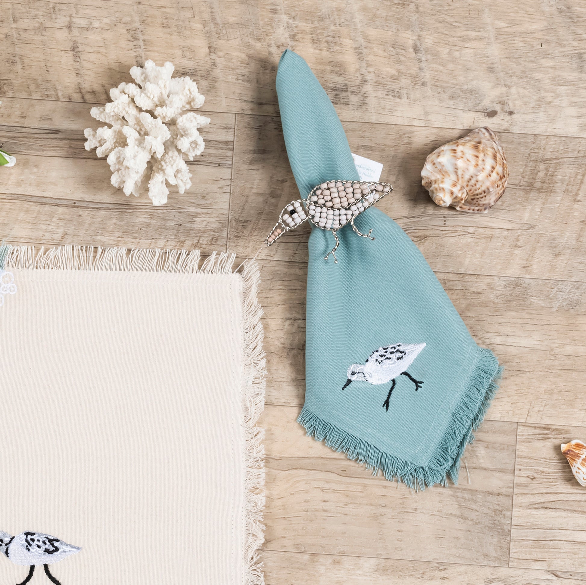 Sandpiper embroidered on coastal blue napkin next to two sandpipers embroidered on a cotton fringed placemat featuring blue waves on sand. Linens are sitting on a wooden table.