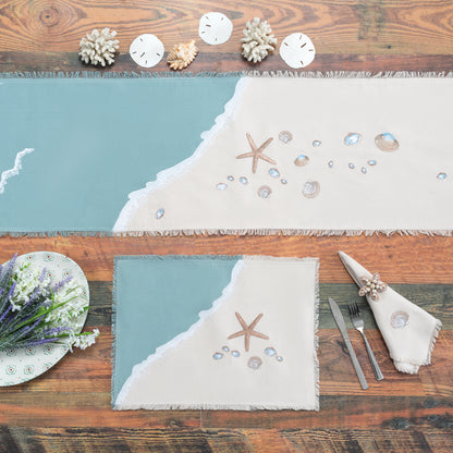 Sea stars and sea shells embroidered on a cotton runner and placemat featuring a wave washing onto the beach, finished with fringe edges. A natural cotton fringed napkin featuring an embroidered scallop shell.