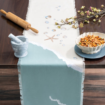 Sea stars and sea shells embroidered on a cotton runner  featuring a wave washing onto the beach, finished with fringe edges on a table with snacks and cooking tools.