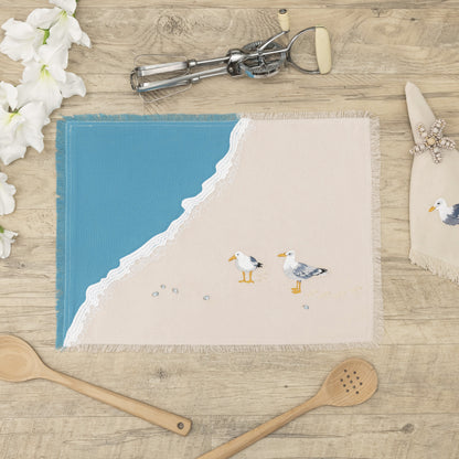 Two sea gulls embroidered on a cotton fringed placemat featuring blue waves on sand. Linens are sitting on a wooden table.