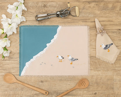 Sea gull embroidered on natural cotton napkin next to two gulls embroidered on a cotton fringed placemat featuring blue waves on sand. Linens are sitting on a wooden table.