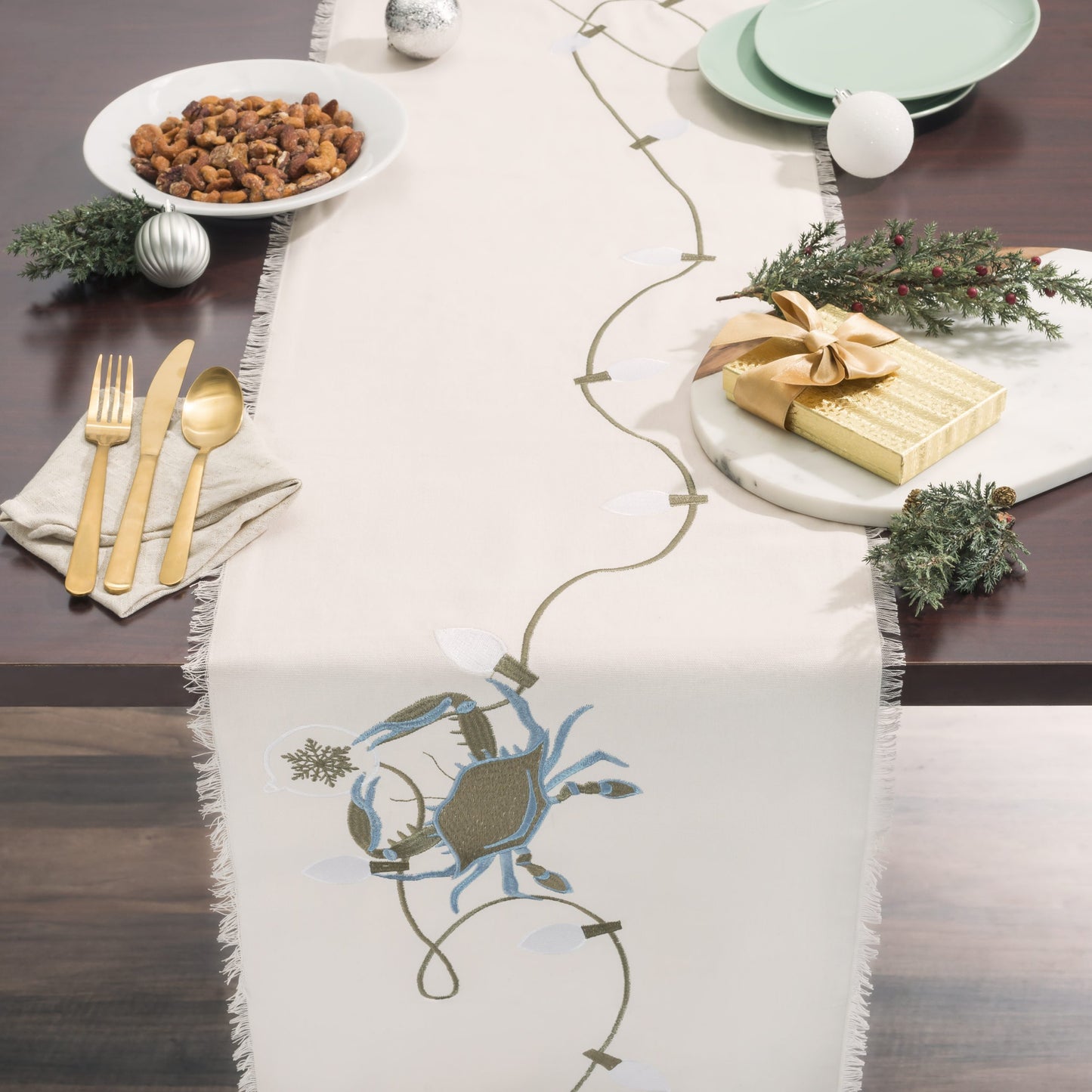 Holiday table setting featuring embroidered table linen collection features a blue crab and holiday lights on natural cotton with fringed edges. 