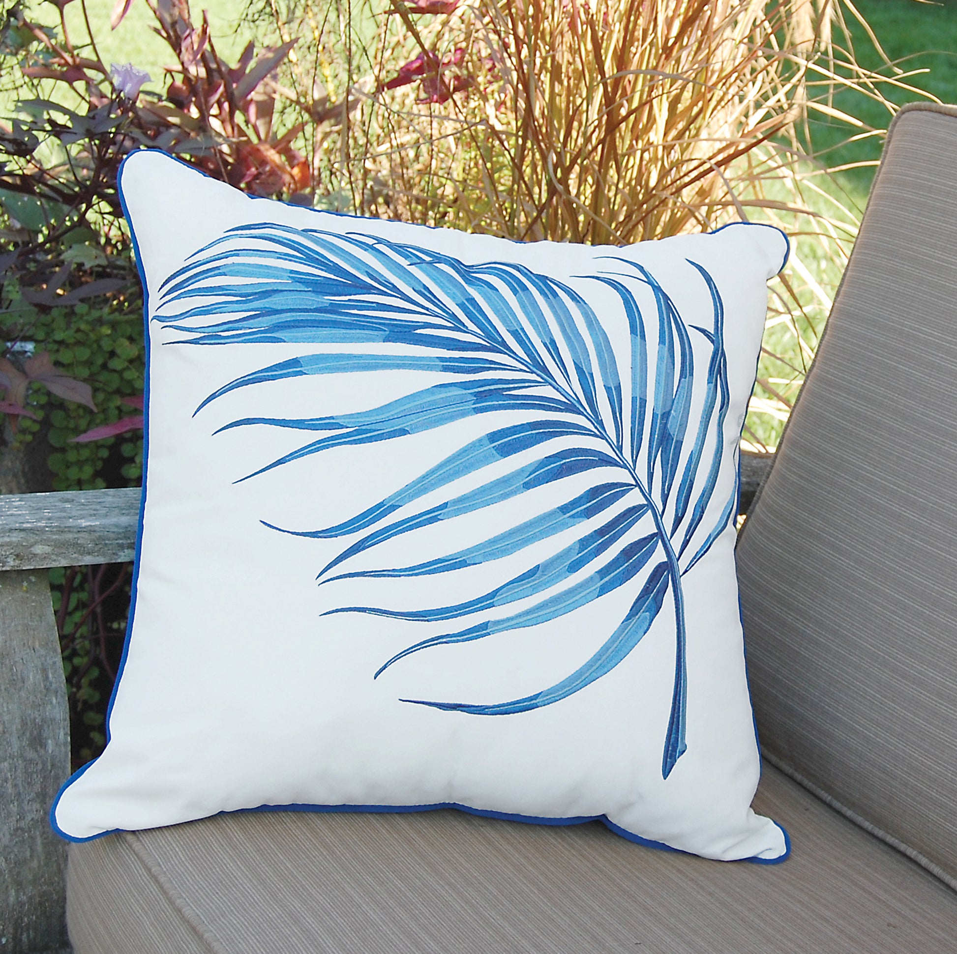 Blue parlor palm outdoor pillow styled on a patio chair next to tall grass.