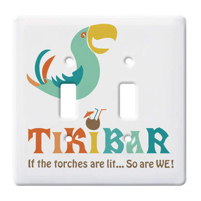 ceramic double toggle switch plate featuring a print of a colorful parrot above a coconut drinking vessel with embellishments and text that reads "tiki bar, if the torches are lit...so are we!"